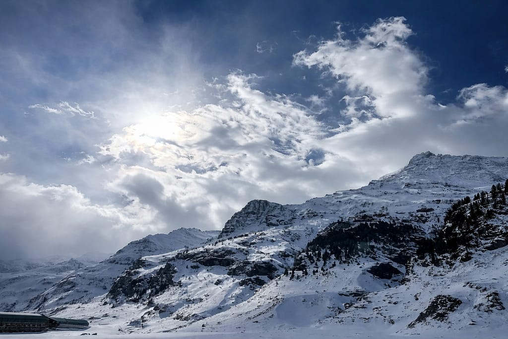 A wide scenic view of snow-capped Himalayan mountains under a bright sun and cloudy sky.